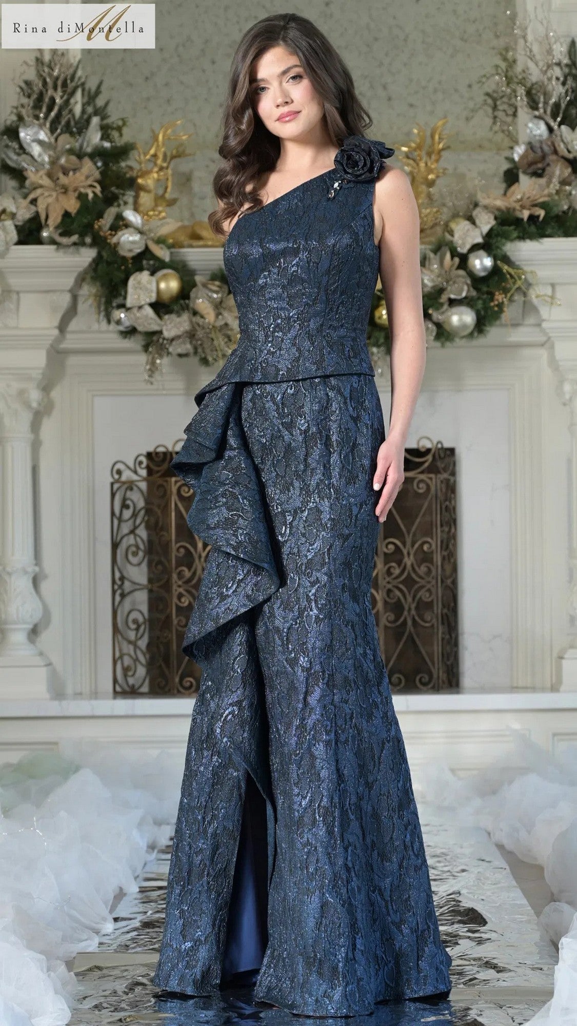 A woman stands indoors wearing the Rina Di Montella RD3231, a navy blue, one-shoulder, floor-length formal dress with floral details and a side ruffle. An ornate fireplace and holiday decorations are in the background.